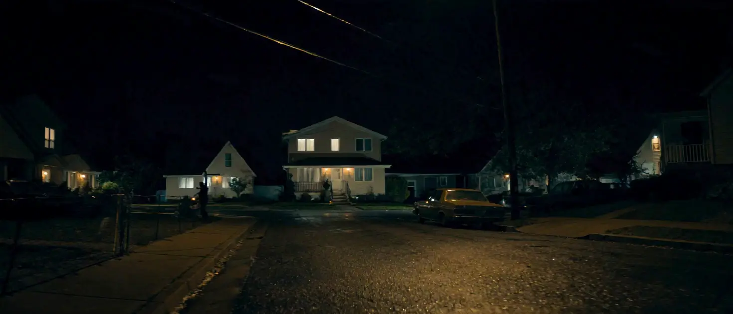 Night view of a residential street: a two-story house with lit windows, a car at the curb, wet asphalt.